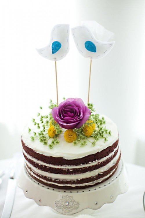 a red velvet wedding cake with frosting, fresh yellow blooms, a pink rose and greenery and fabric bird cake toppers is a creative DIY idea for a relaxed wedding