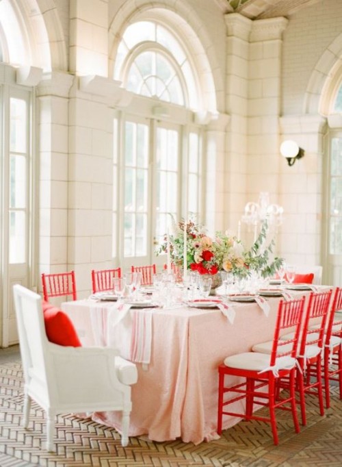 a chic Valentine wedding reception space with lush floral with neutral and red blooms and greenery, a pink tablecloth and red chairs is a lovely and romantic space