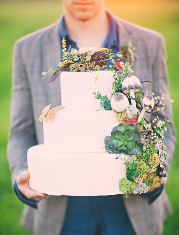 A pure white wedding cake topped with moss, succulents, faux mushrooms, a butterfly and even a faux nest on top