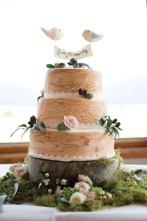 A woodland wedding cake that imitates wood, with fresh blooms and greenery and a bird topper with a banner