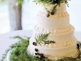 a chic white wedding cake topped with pinecones and ferns, on a elegant stand covered with evergreens