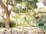 an under the trees wedding reception space with tables styled with pink and neutral blooms and accented with greenery pendants and woven pendant lamps
