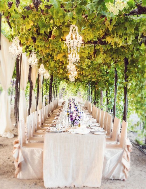 a sophisticated vintage vineyard wedding reception with greenery around, neutral curtains, neutral linens, neutral chair covers and crystal chandeliers