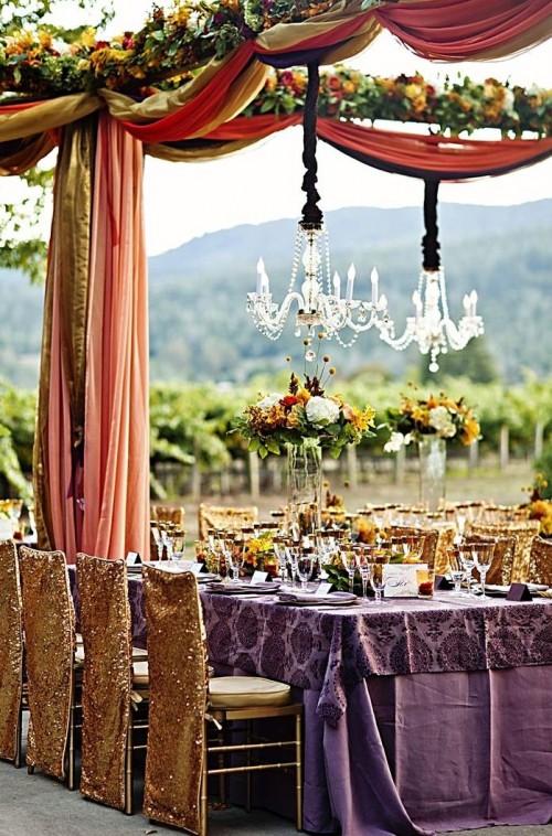 a colorful and sophisticated vineyard wedding reception with yellow and orange fabric, with bold blooms and greenery, a purple tablecloth and gold sequin chair covers plus crystal chandeliers