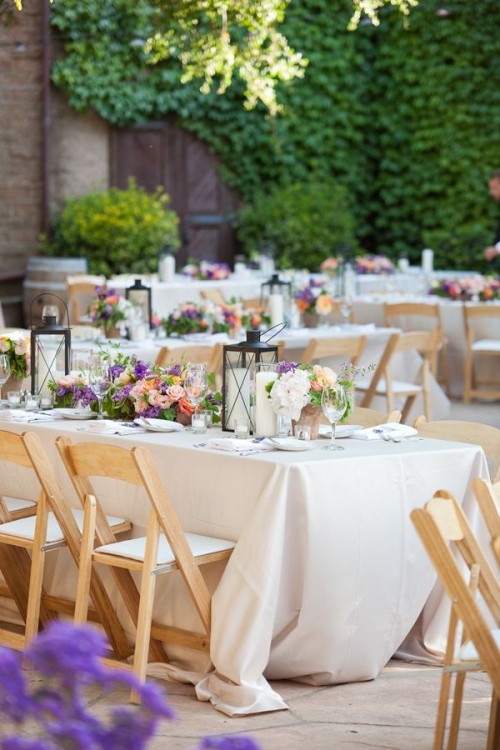 a vineyard wedding reception with neutral tablecloths, bright blooms and greenery and candle lanterns and greenery around the space