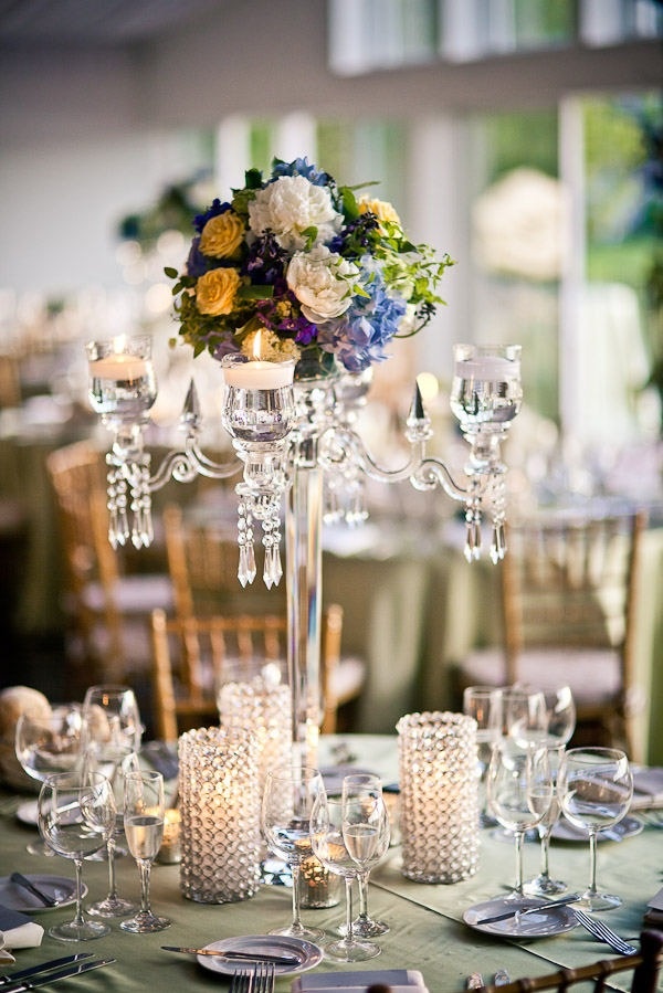 a refined summer wedding centerpiece of a crystal candelabra with candles and bright blooms and greenery and with candles in crystal candleholders