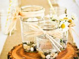 a simple summer wedding centerpiece of a wood slice, jars with pebbles, floating candles and daisies for a relaxed woodland summer wedding