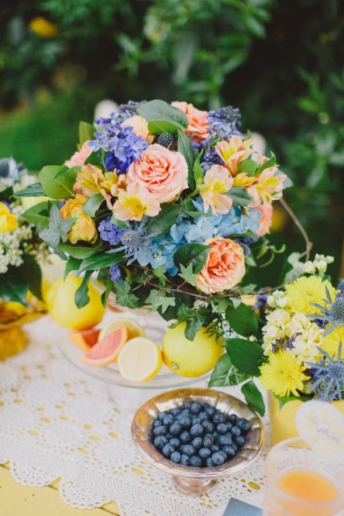 a vivid summer wedding centerpiece of yellow, blush, blue blooms, thistles and leaves and citrus is summer-inspiring