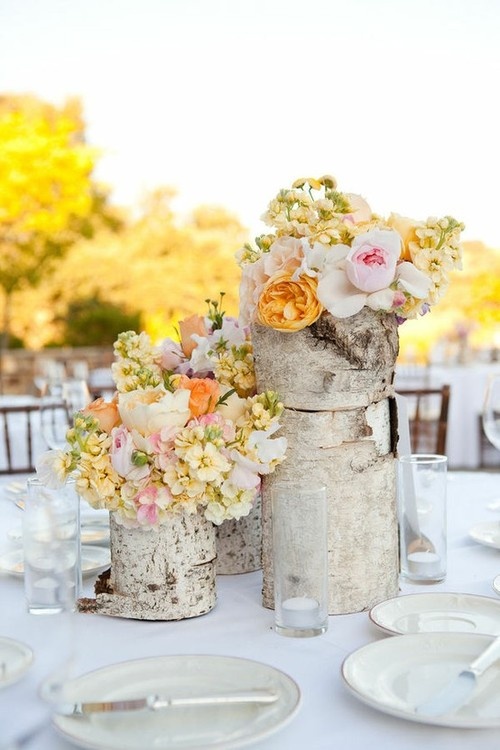a pastel summer wedding centerpiece of tree stumps, peachy, blush, white and yellow blooms, candles in tall glasses