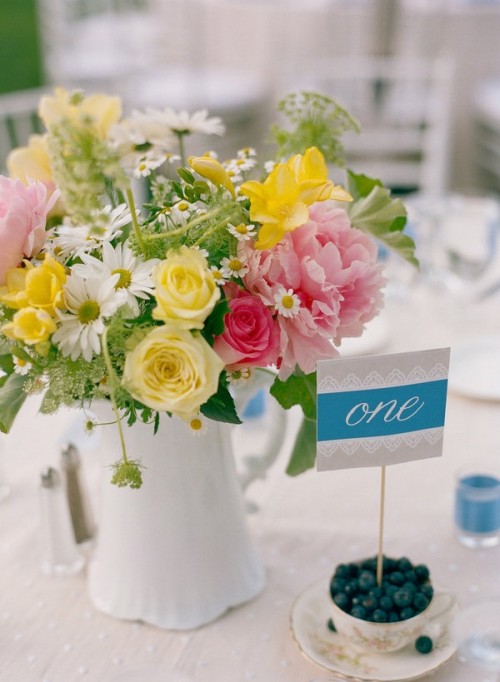 a vibrant summer wedding centerpiece of a white jug, yellow, pink and white blooms and greenery and a card on a stand