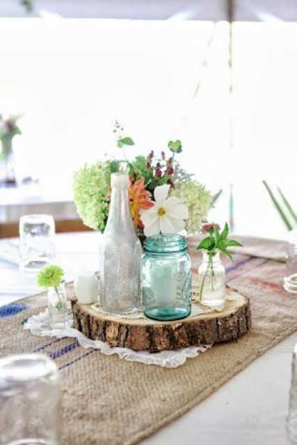 a rustic summer wedding centerpiece of a wood slice, neutral and green blooms and bottles and jars