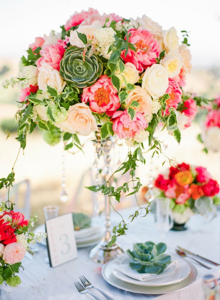 a colorful refined summer wedding centerpiece of a silver stand, white, blush, pink blooms, succulents and cascading greenery is very vibrant