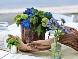 a driftwood wedding centerpiece with green and purple blooms, thistles and some candles for a beach or coastal wedding