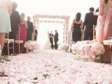 a wedding arch fully covered with pink roses and peony roses and a wedding aisle fully covered with their petals for a romantic feel