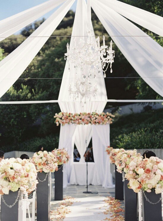 a fabulous wedding ceremony space done with white curtains, a chic crystal chandelier, neutral and pink floral arrangements and crystals is wow