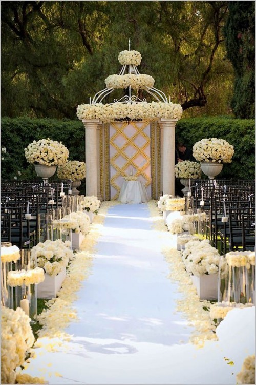 an exquisite wedding ceremony space done with lots of white rose arrangements and a wedding backdrop covered with white roses on top