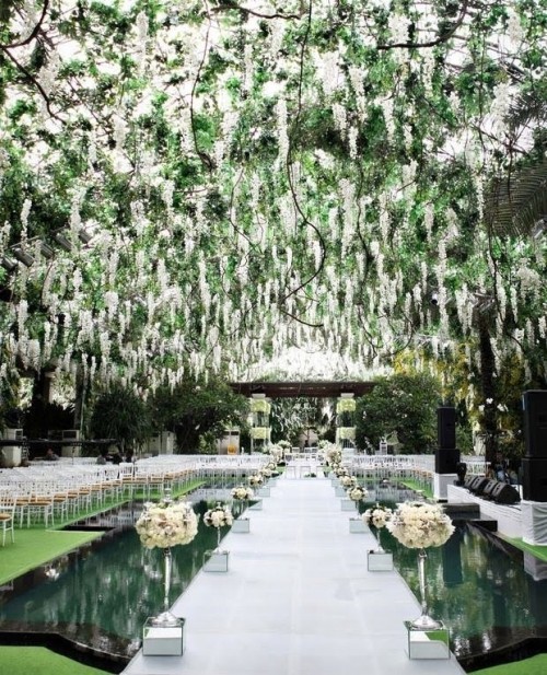 a jaw-dropping wedding ceremony space wiht greenery and blooming branches hanging over it, white flower arrangements lining up the aisle