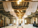 a barn wedding ceremony space with white petals on the floor, white curtains on the ceiling and white chairs is a pretty and inspiring space