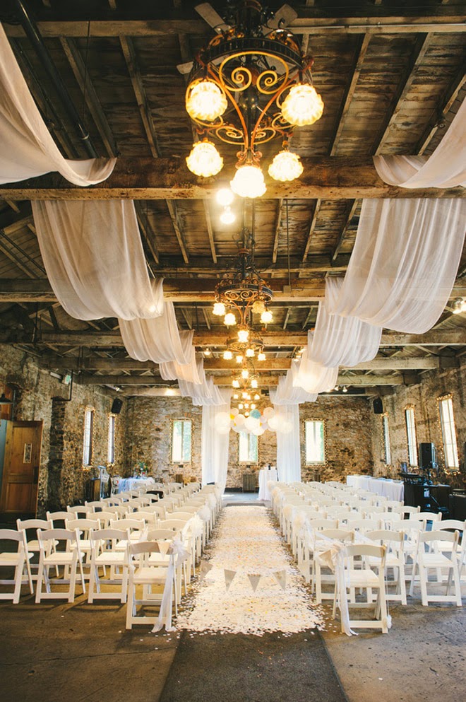 A barn wedding ceremony space with white petals on the floor, white curtains on the ceiling and white chairs is a pretty and inspiring space