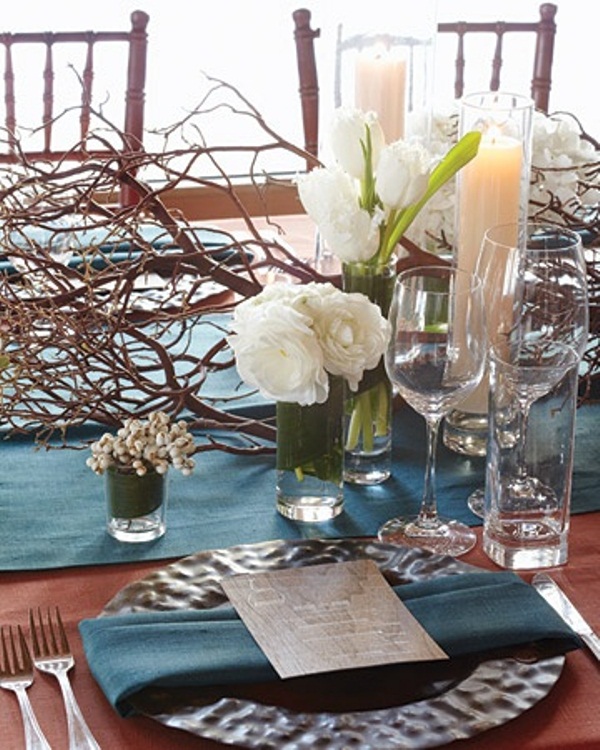 a winter wedding tablescape with a blue fabric table runner, napkins, a hammered charger, white blooms and a branch decoration