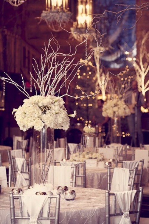 a white and silver winter tablescape with silver candleholders, a tall clear vase with branches and white blooms