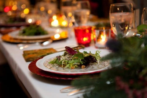 a green and red winter wedding table with lots of lights and candles in candleholders, evergreens and silver cutlery