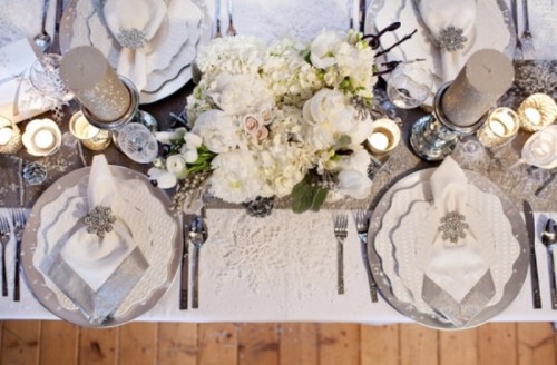 a silver, grey and white winter wedding table setting with grey sparkling candles, chargers, a shiny runner, white blooms and silver cutlery