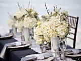 stylish black, white and silver winter wedding tablescape with white floral centerpieces, sivler napkins with white napkin rings, mercury glass candleholders and a black tablecloth