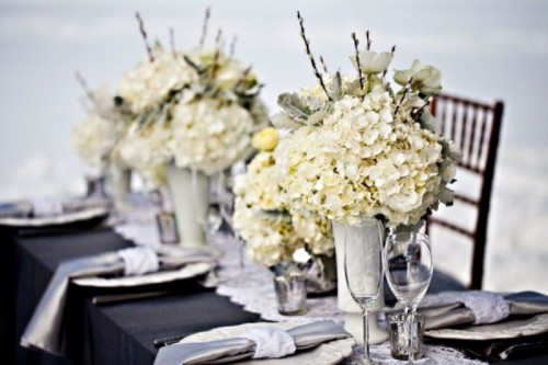 stylish black, white and silver winter wedding tablescape with white floral centerpieces, sivler napkins with white napkin rings, mercury glass candleholders and a black tablecloth